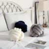 Three knot ball cushions in cream, grey, and navy displayed on a white bed in a cozy, minimalistic bedroom.