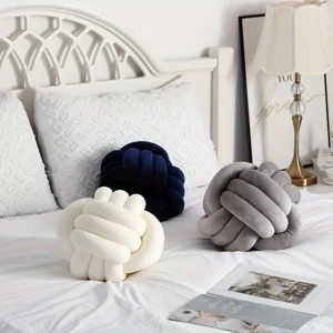 Three knot ball cushions in cream, grey, and navy displayed on a white bed in a cozy, minimalistic bedroom.