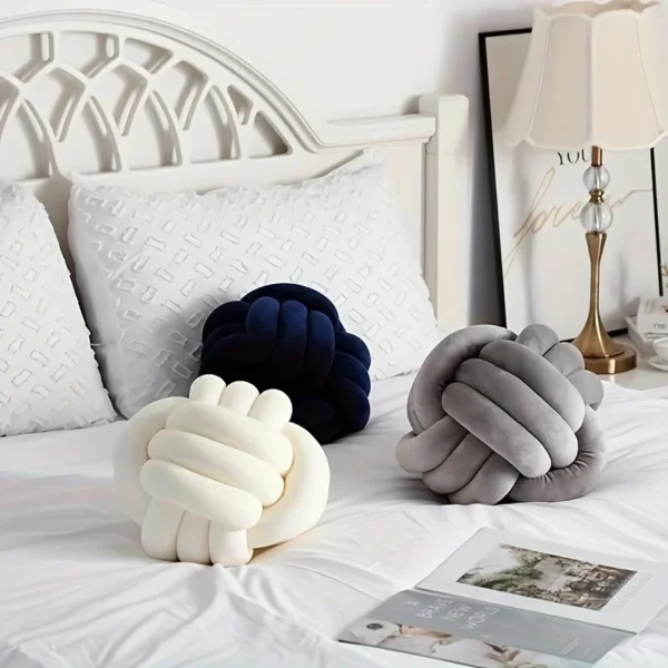 Three knot ball cushions in cream, grey, and navy displayed on a white bed in a cozy, minimalistic bedroom.